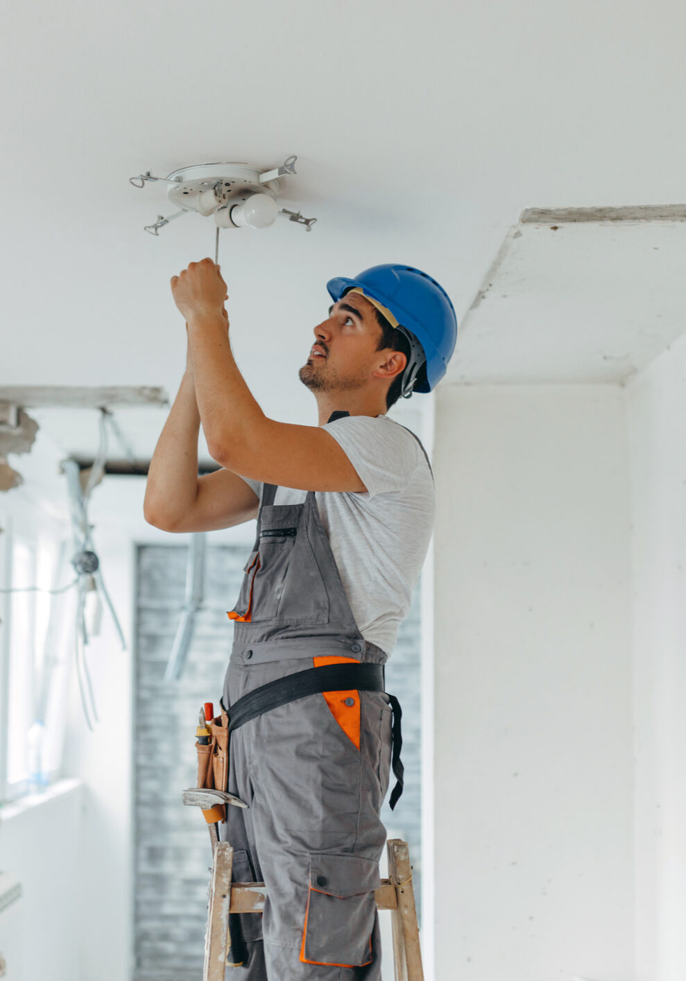 A young Caucasian male construction worker is using a screwdriver to fix a light bulb, while standing on a ladder.